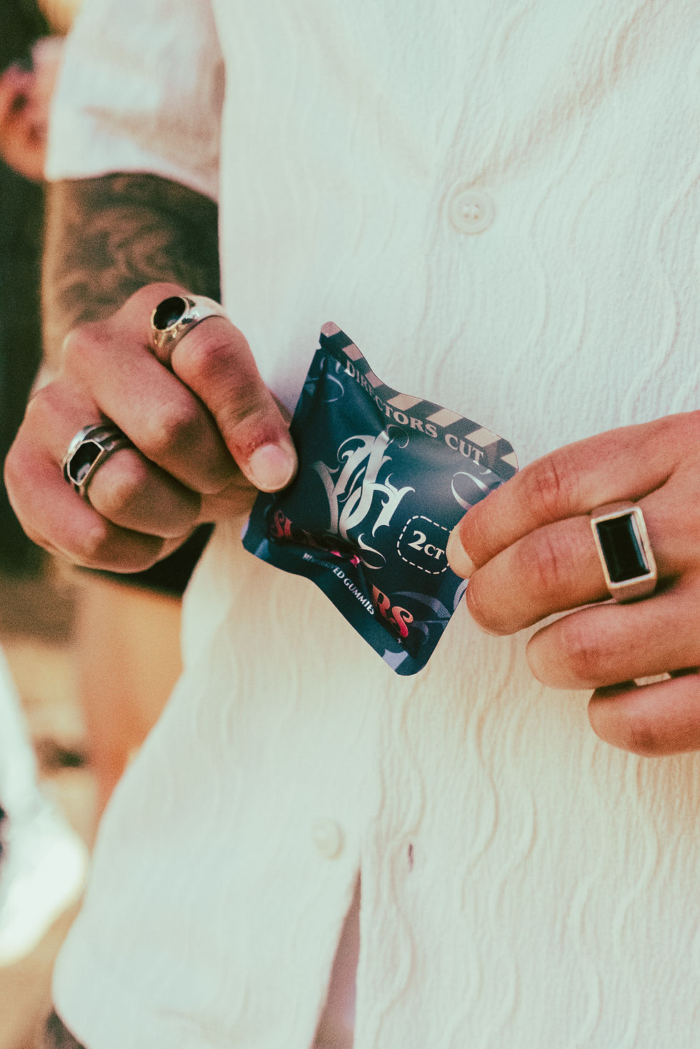 Hands holding a blue packet labeled "2 ct" with tattoos and rings visible. Person wears a textured white shirt. Warm outdoor setting.