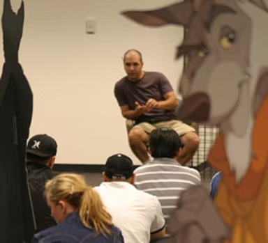 Len sitting in front of a group, talking, beside a large painted cut-out of an animated character