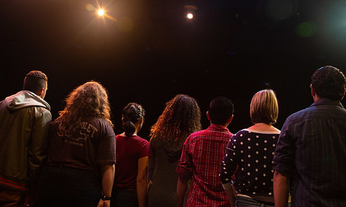 The backsides of seven young performers of varying ethnicities onstage together in a production of 'Untold'