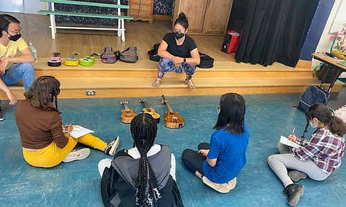 A teaching artist with a black face mask sits in a circle with a small group of young students taking notes during a school tour workshop