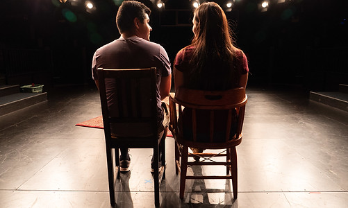 The backsides of two young actors onstage together seated in different wooden chairs during a performance of 'Qulili'