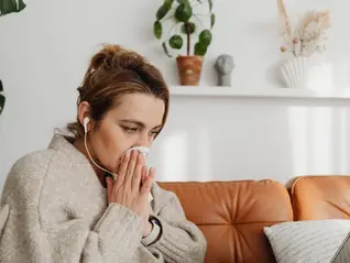 Woman in sweater sits on a brown sofa, using a tissue while wearing earphones. Plants on a white shelf in the background. Cozy atmosphere.