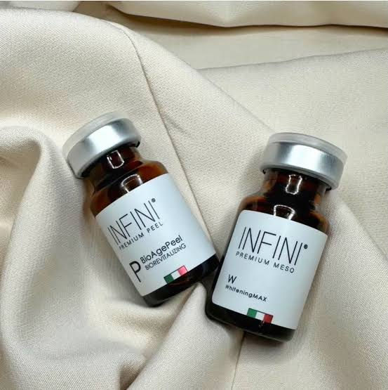 Two brown bottles labeled INFINI on beige fabric. One reads BioAgePeel, the other WhiteningMAX, with Italian flag colors on labels.