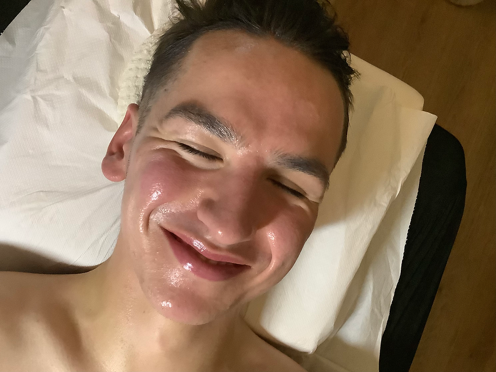 Smiling person with closed eyes, lying on a white sheet, face shiny and relaxed, wooden background adds warmth to the image.