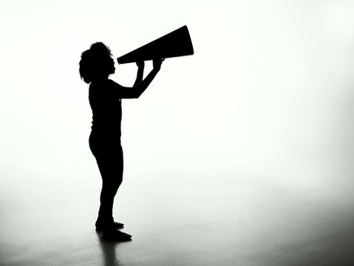 A silhouette of a person with curly hair standing in profile, holding a megaphone up to their mouth with both hands. The figure appears to be shouting or making an announcement through the megaphone. The image is rendered in black silhouette against a light gray gradient background, creating a dramatic contrast that emphasizes the act of communication or protest.