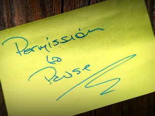 A yellow sticky note with blue handwriting that reads, “Permission to Pause,” placed on a wooden surface.