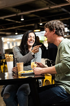 A woman and man sitting at a table laughing, while she feeds him a piece of a waffle.