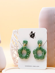 green daisy shaped earrings hanging from a smaller square stud earring. The surface features a white leaf pattern and is presented in a high gloss finish. Gold coloured stainless steel findings finish the earrings.