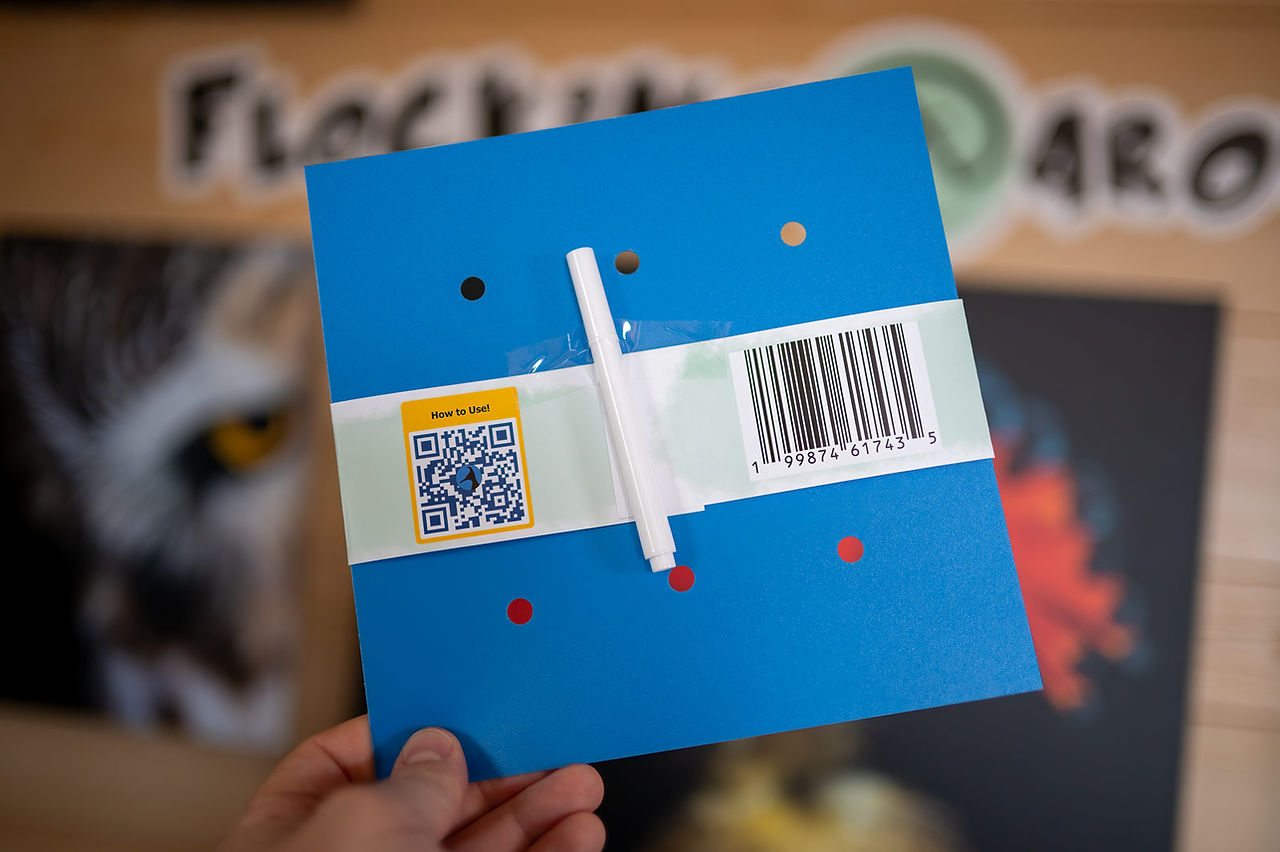 Blue "Flock Blocker" held in hand, viewing the back with a QR code and UPC, aims to protect birds from windows. Background shows part of a sign with bird imagery. Mood is informative.
