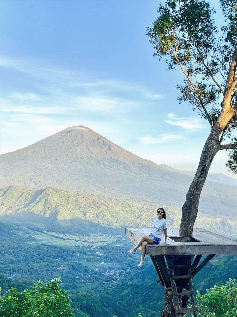 Woman in sunglasses sits on a tree platform above a green valley, with a volcano and blue sky in the background.