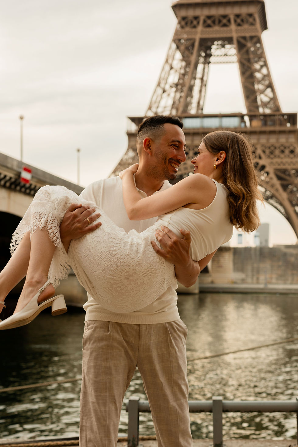 Man in white shirt holds woman in lace dress by Eiffel Tower, both smiling against a river backdrop under a cloudy sky. Romantic mood.
