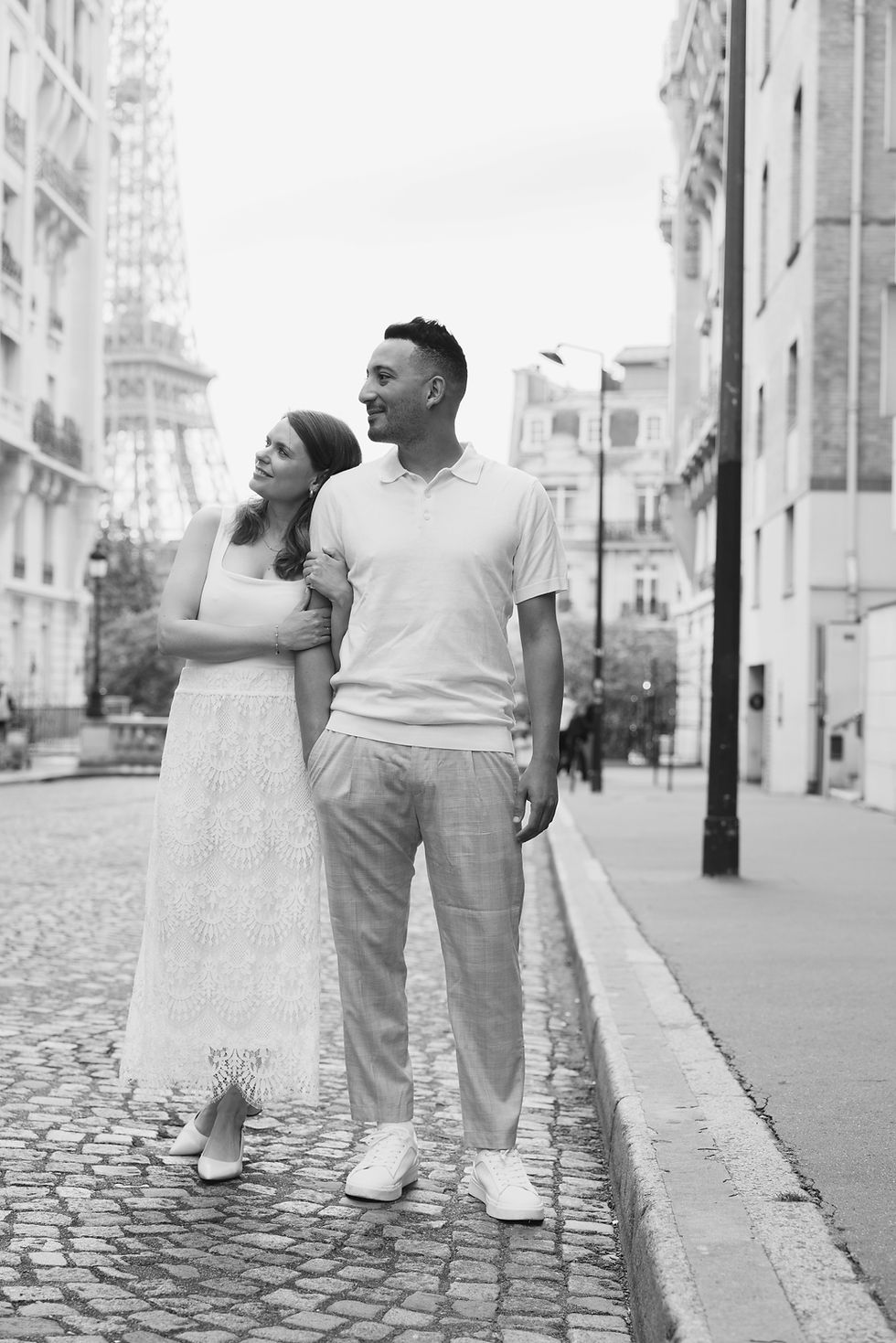 A smiling couple in casual white attire walks on Avenue des Camoens street with the Eiffel Tower in the background, conveying a romantic mood.