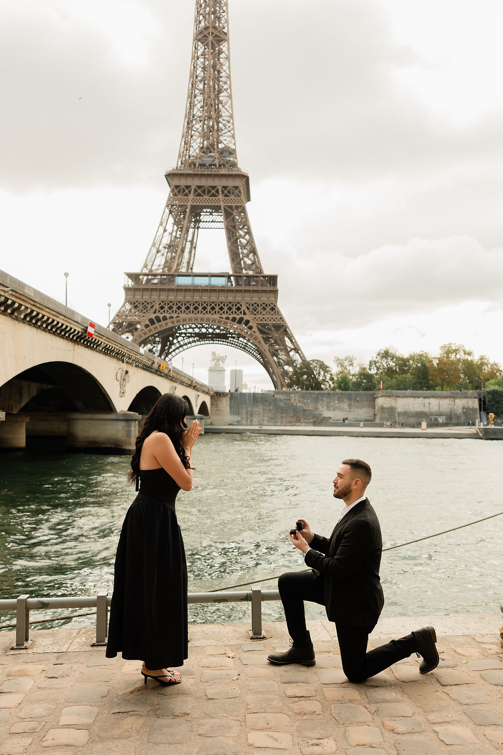 Man kneels by the Seine and proposes to a surprised woman, with the Eiffel Tower behind them under a cloudy sky.