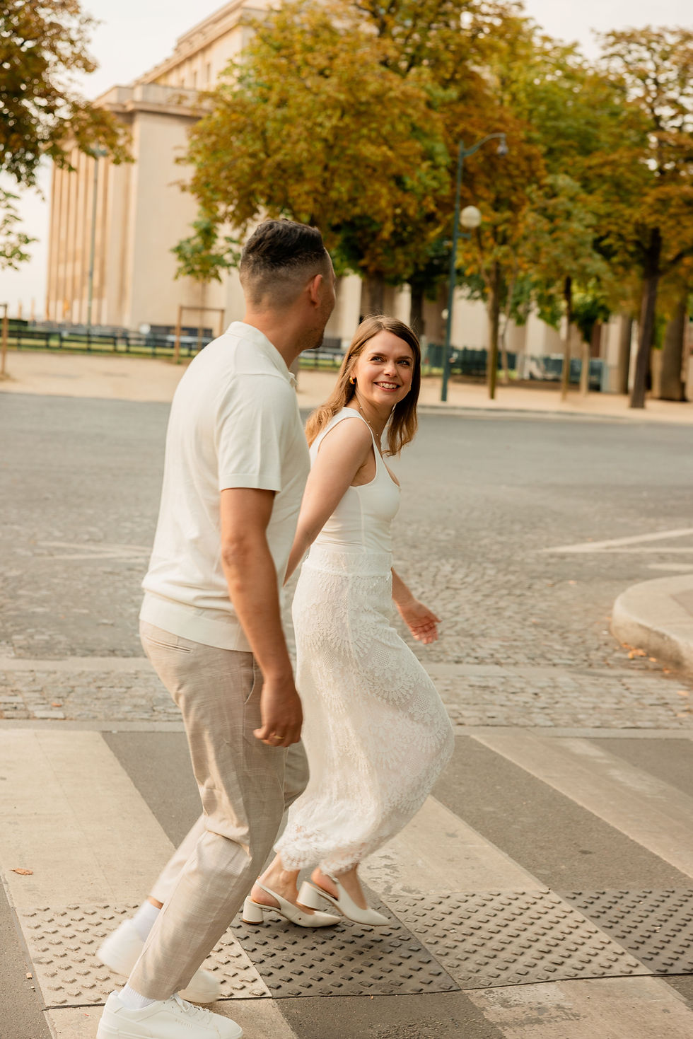 Couple walks hand in hand on Trocadero city streets, surrounded by trees and buildings, wearing white outfits. The setting is sunny and cheerful.