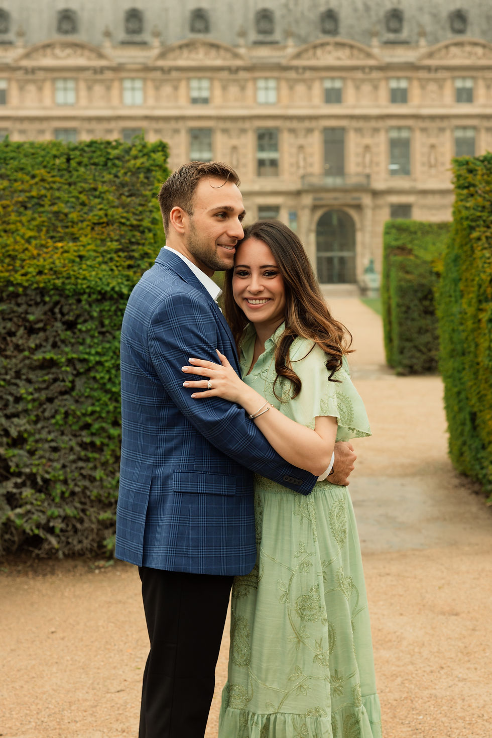 Smiling couple embracing in a formal garden, with a grand stone mansion and trimmed hedges behind them.