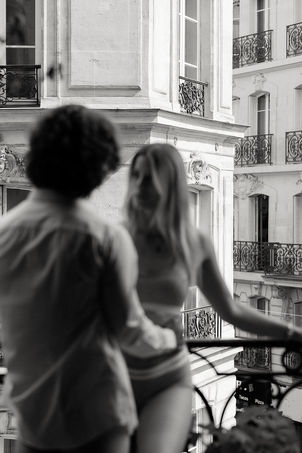 Two people in intimate conversation on a balcony overlook Haussmannian Parisian buildings. Monochrome setting with wrought iron railings; romantic mood.