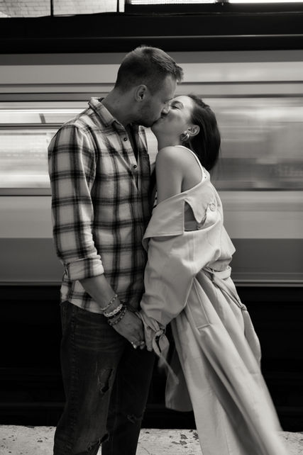 A couple kisses in front of a motion blurred metro. Photo is black and white.