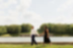 A couple walks hand in hand near a reflective pond, surrounded by green trees under a partly cloudy sky, conveying a serene mood.