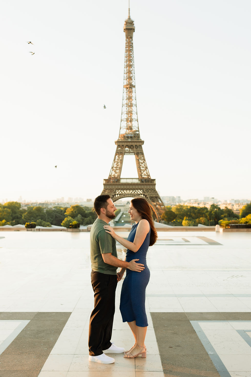 Smiling couple embraces on a terrace with the Eiffel Tower behind them at sunset.