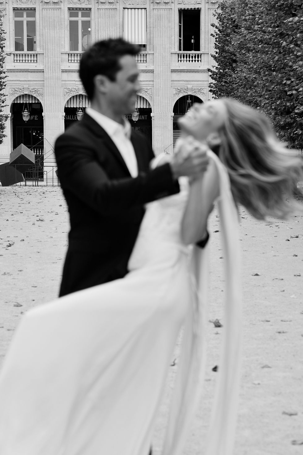 A couple dances joyfully outside a Palais Royale, her white dress flowing. Blurred motion captures their emotion. Trees and columns in view.