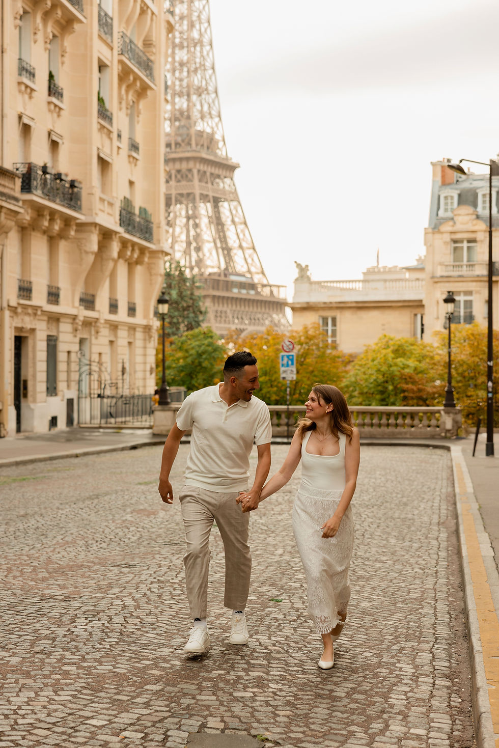 Couple holding hands, walking on a cobblestone street near the Eiffel Tower. Soft light, beige buildings, and green foliage. Romantic mood.