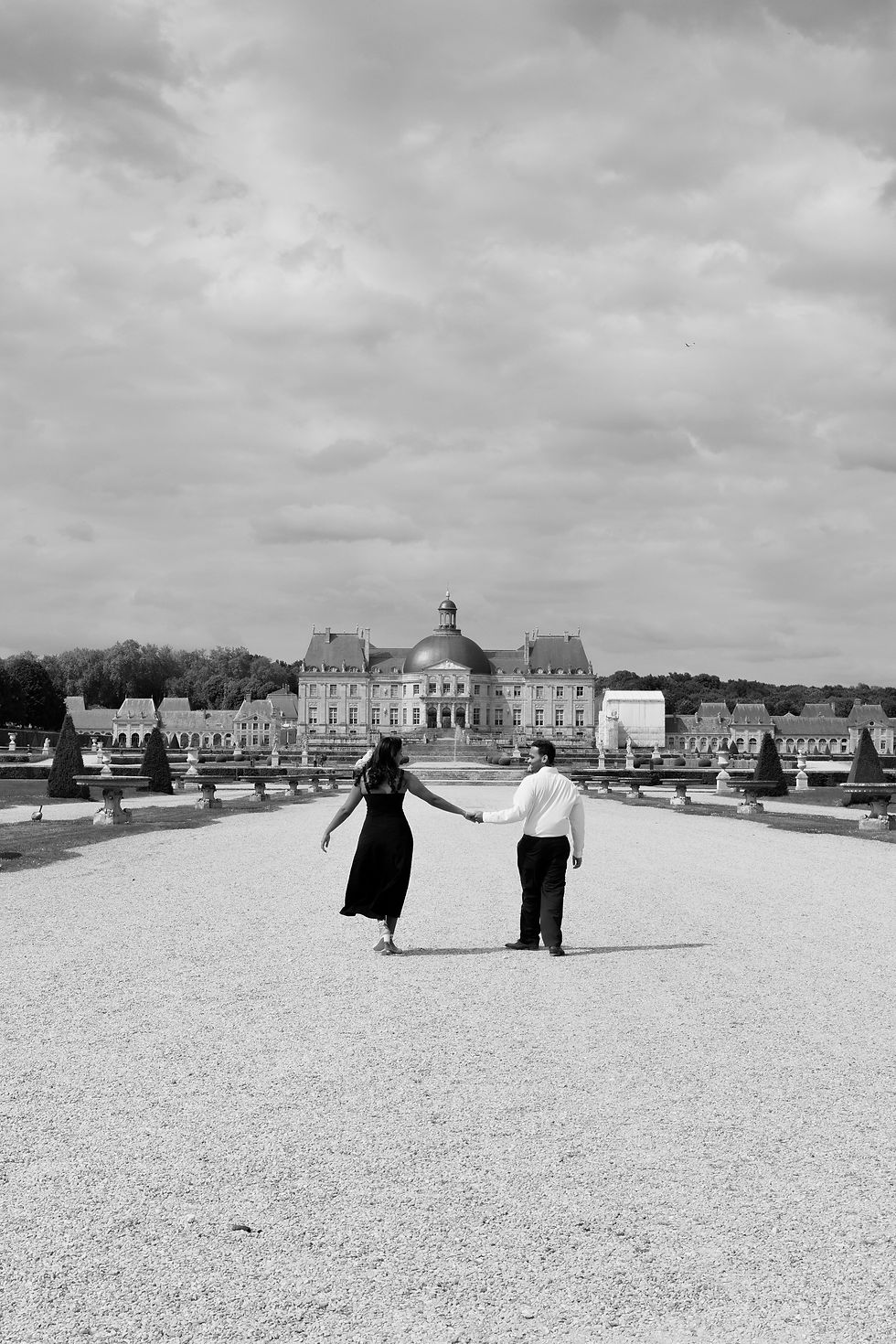 A couple holding hands walks on a gravel path towards a grand chateau under a cloudy sky. The scene conveys romance and elegance.