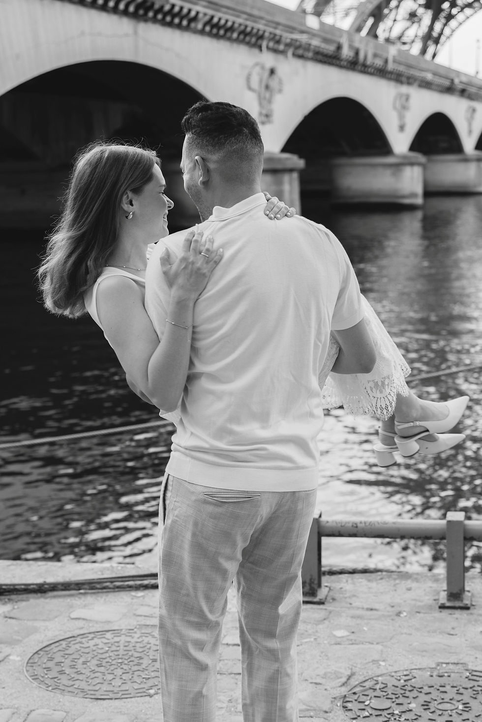 Man carrying a woman by the Seine. They smile at each other with a bridge in the background. Black and white, romantic mood.