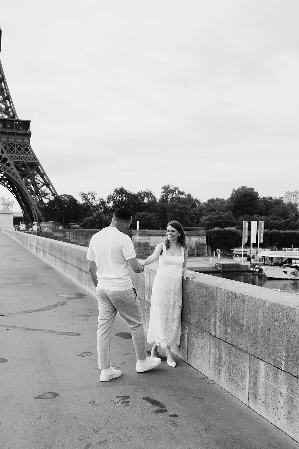 A couple stands by a stone railing near the Eiffel Tower. The man, in white, reaches out to the woman, who smiles wearing a white dress.
