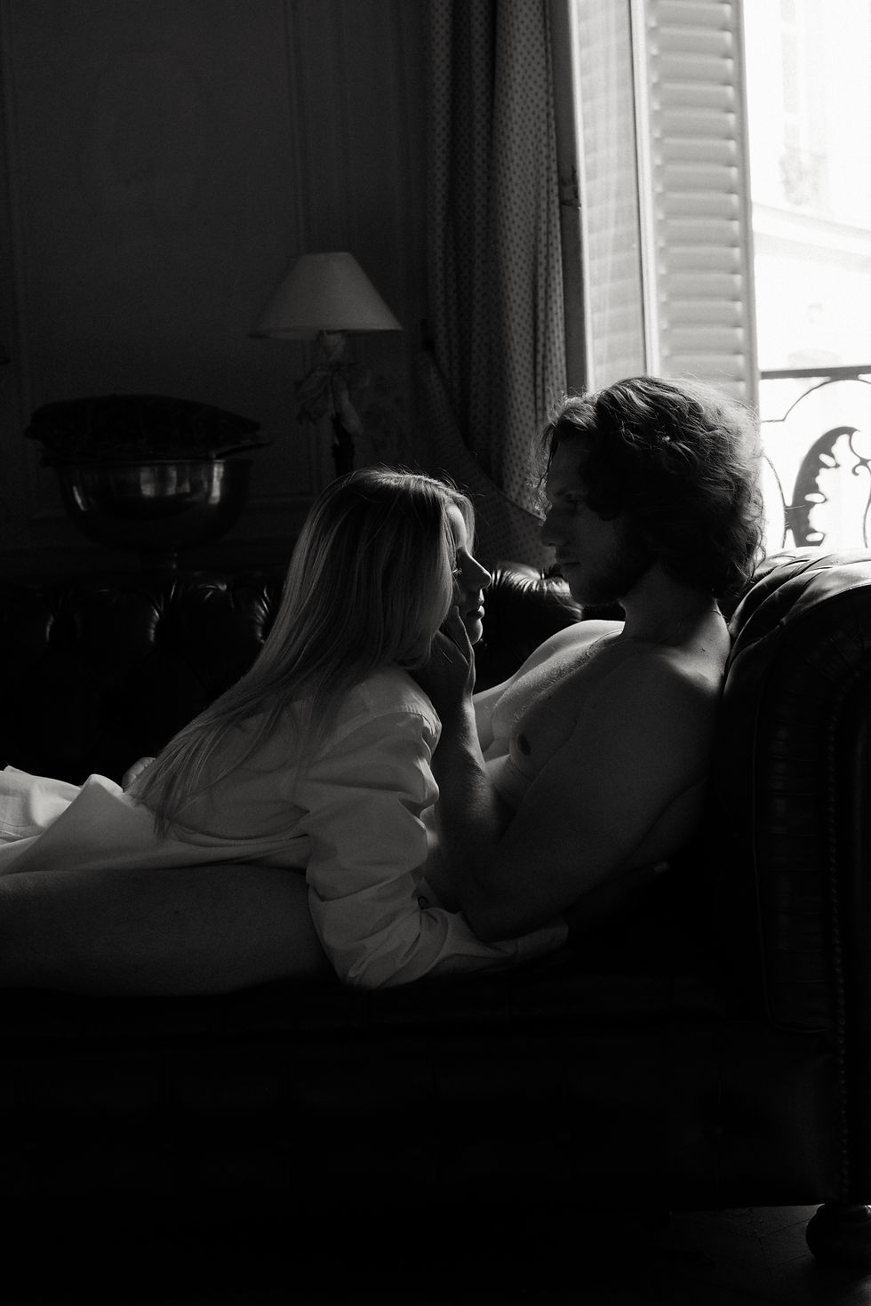 A couple embraces intimately on a sofa in a dimly lit room. A lamp and open window in the background add a romantic, cinematic, and serene mood.