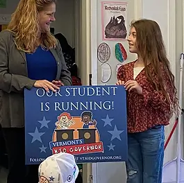A blond haired woman in a green button down shirt presents a young girl with long brown hair and a burgundy flowered blouse with a sign that reads "Our Student is Running"