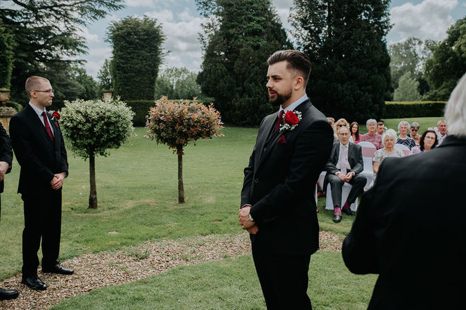 Groom in black suit with burgundy tie and buttonhole waits nervously for his bride to arrive.