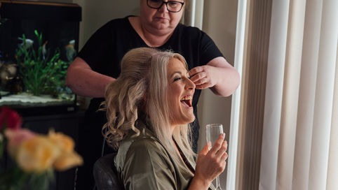 A bride sipping champagne and laughing as she is having her hair styled for her wedding in Northamptonshire