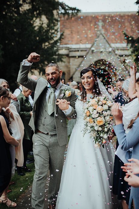 A bride and groom getting covered in confetti on their wedding day in bedfordshire captured by Tara Gillen Photography. The bride is carrying a beautiful wedding bouquet of neutral peach and white roses