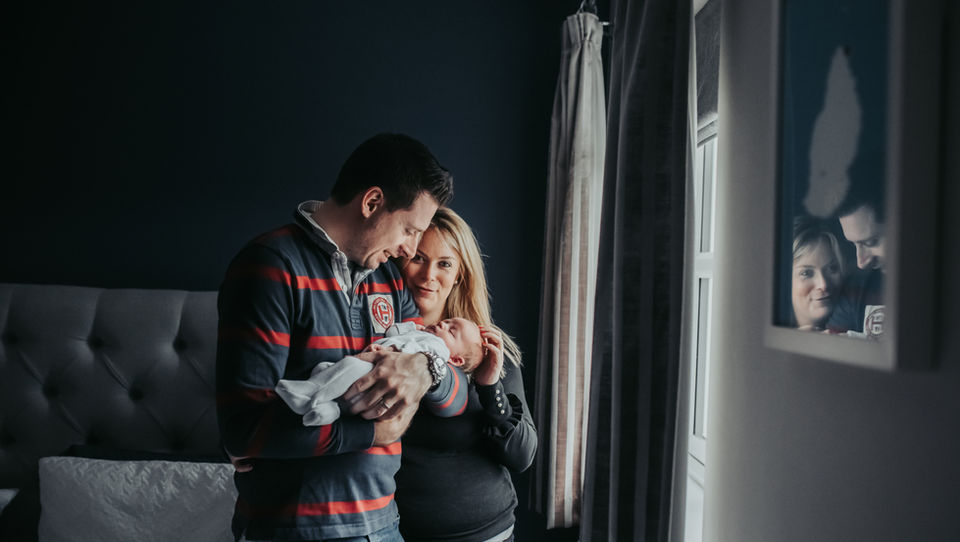 Parents cradle their newborn baby as they stand relaxed beside a window at their home in Bedfordshire captured by Tara Gillen Newborn Photographer