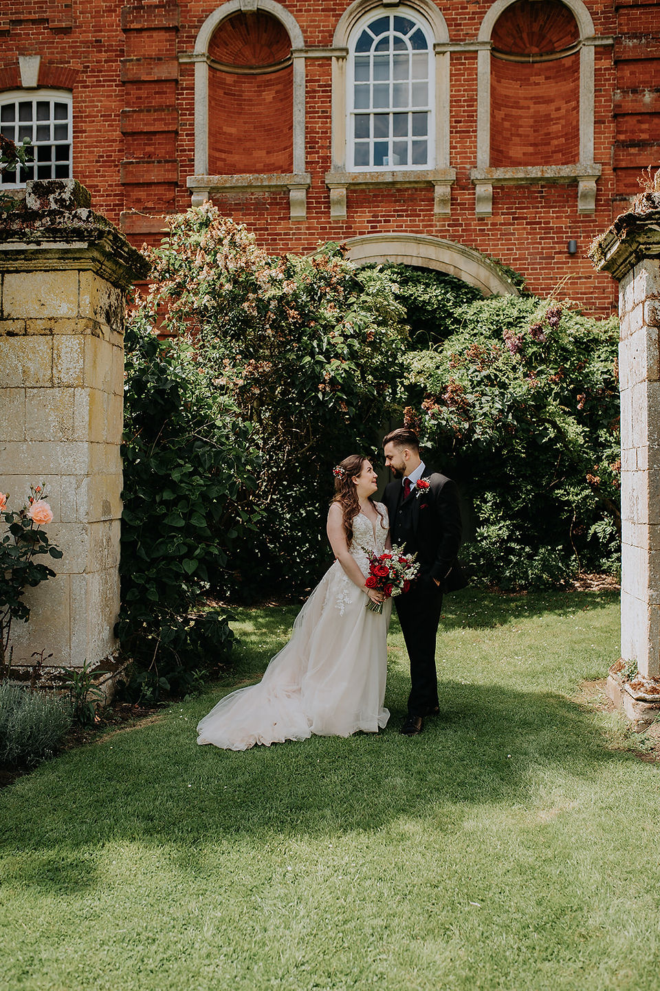 Groom in a black suite with burgundy buttonhole stares lovingly at bride stood beside him.