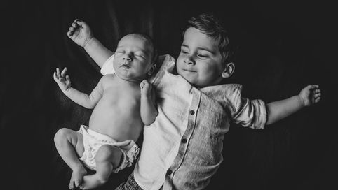 A black and white photo of a young boy lying down and relaxing with his newborn sibling during their photoshoot with Tara Gillen Photography at their home in Bedford