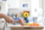 Hands chop carrots on a wooden board in a bright kitchen. There's a bouquet with a sunflower in the background, adding a cheerful touch.