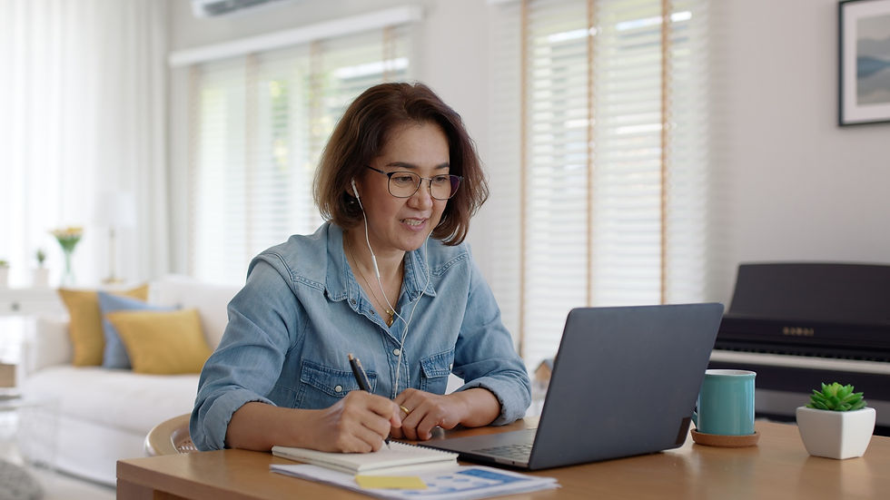 Woman in glasses smiling while writing in a notebook and looking at a laptop. She's wearing earphones. Cozy room with sofa, lamp, and plants.