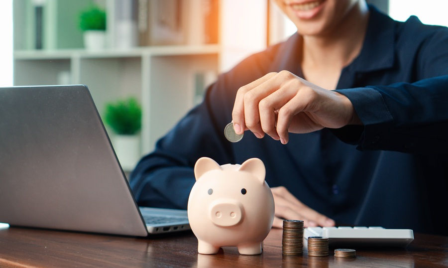 Smiling person placing coin into a piggy bank on a desk with a laptop, stacked coins, and a keyboard in a bright office setting.