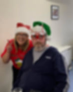 Two people in festive hats; woman smiling and posing with peace sign, man in a green hat. They're indoors with a photo on the wall.