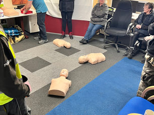 CPR training in a room with mannequins on a patterned carpet. People watch a man preparing equipment. Casual setting with relaxed mood.