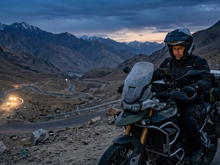 A atmospheric twilight photograph on a winding mountain road in Ladakh, India. A group ride leader on a heavily equipped adventure motorcycle is stopped safely at a road shoulder overlooking a curving pass. He is looking at his handlebar-mounted smartphone displaying the RidingVerse dark-mode app interface showing the 'Live Group Tracking' map with multiple real-time rider pins scattered along the route behind him. In the background down the valley, the headlights of three approaching group members illuminate the dusk haze