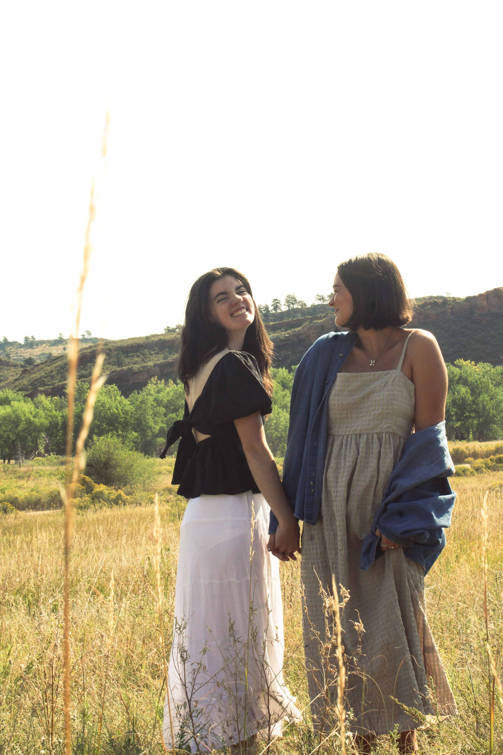 Two girls holding hands, one looking at the other while she is looking at the camera.