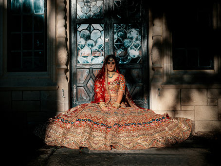 A woman in a vibrant traditional Indian lehenga sits elegantly in front of an ornate door, illuminated by soft lighting, creating a regal and festive ambiance.