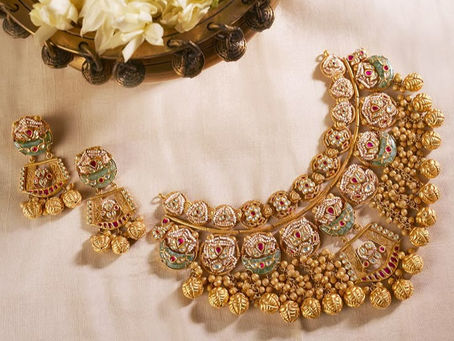 A set of traditional Indian jewelry, including an intricately designed gold necklace and matching earrings, adorned with colorful gemstones, placed on a white fabric background.