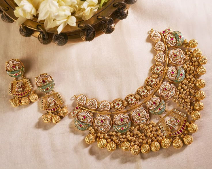A set of traditional Indian jewelry, including an intricately designed gold necklace and matching earrings, adorned with colorful gemstones, placed on a white fabric background.