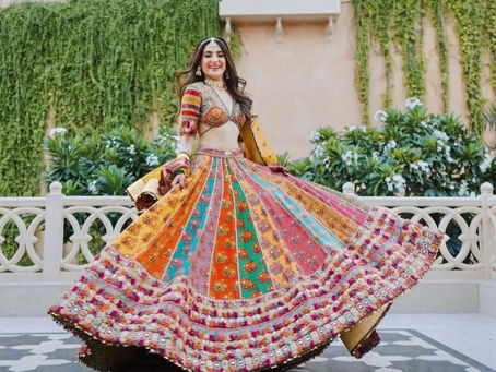 A woman twirling in a vibrant, multicolored lehenga with intricate embroidery, standing in an elegant outdoor setting with greenery and white architectural elements.