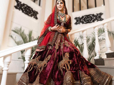A woman wearing a richly embroidered traditional lehenga in deep red and gold, posing elegantly on a grand staircase with lush greenery in the background.