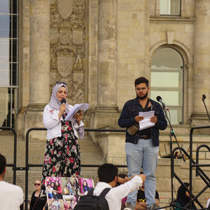 A woman speaks into a microphone, her voice firm with emotion, addressing the crowd of demonstrators gathered beneath grey skies.