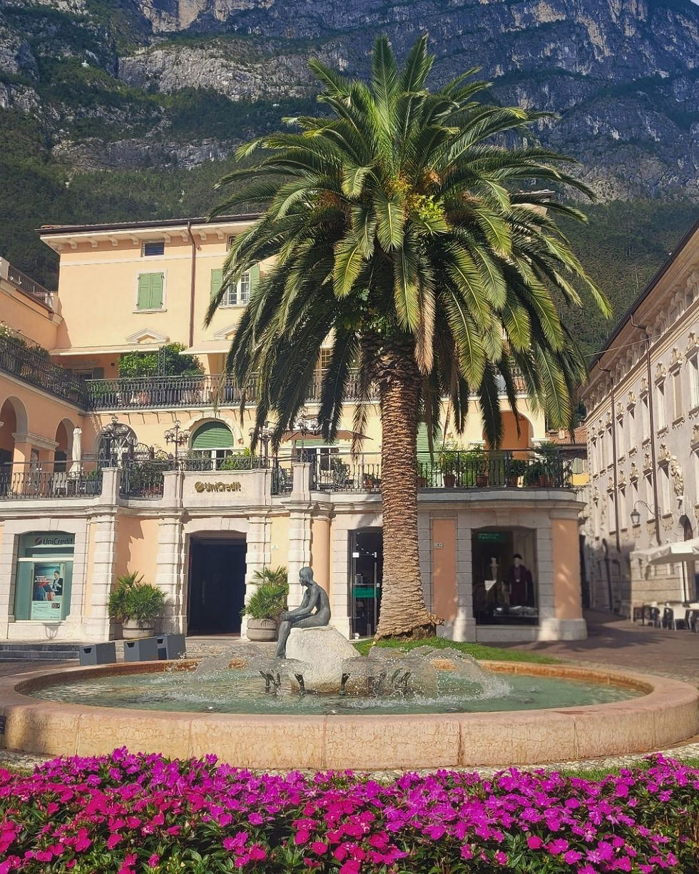 Palm tree and fountain with seated statue in a sunny plaza, pastel buildings, purple flowers, and a UniCredit sign beneath mountains.